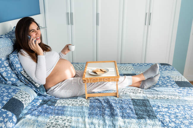 Side view high angle of cheerful pregnant female sitting on bed in morning and having breakfast while speaking on smartphone — Stock Photo