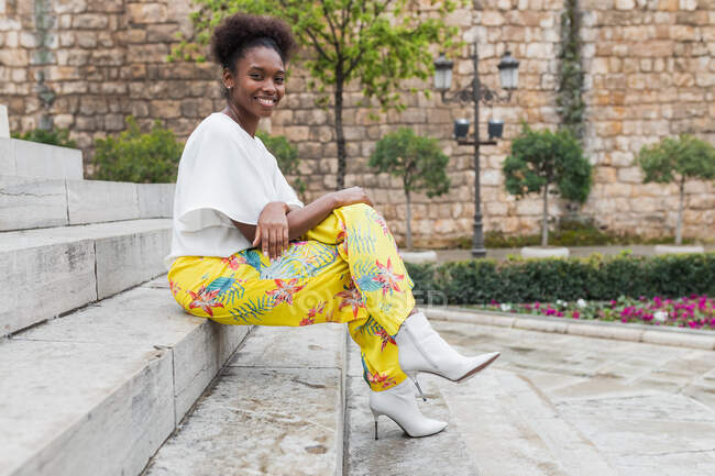Side view full length smiling African American female in trendy outfit sitting with crossed legs on wooden stairs and looking at camera in sunny summer garden — Stock Photo