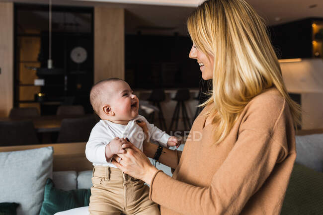 Vista lateral de la madre encantada sosteniendo adorable bebé sonriente mientras se divierten juntos en casa - foto de stock