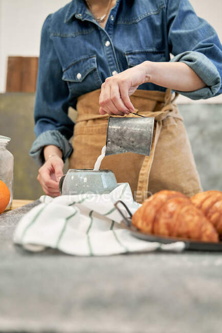 Young female blogger pouring milk from jug into cup against photo camera at table with croissants at home — Stock Photo