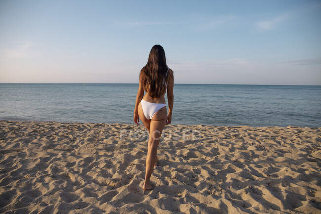 Обратный вид на неузнаваемую женщину в купальниках, идущую по песчаному берегу к океану под облачно-голубым небом — стоковое фото