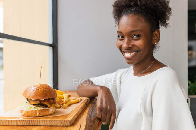 Donna afroamericana che mangia deliziose patatine fritte e gustosi hamburger serviti su tavola di legno su un tavolo alto nel ristorante fast food mentre guarda la fotocamera — Foto stock