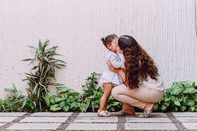 Side view of tender mother kissing cute little child in cheek while standing in yard with green plants — Stock Photo