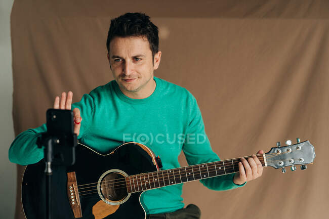 Cheerful adult male vlogger with guitar and plectrum recording video tutorial on cellphone at home — Stock Photo