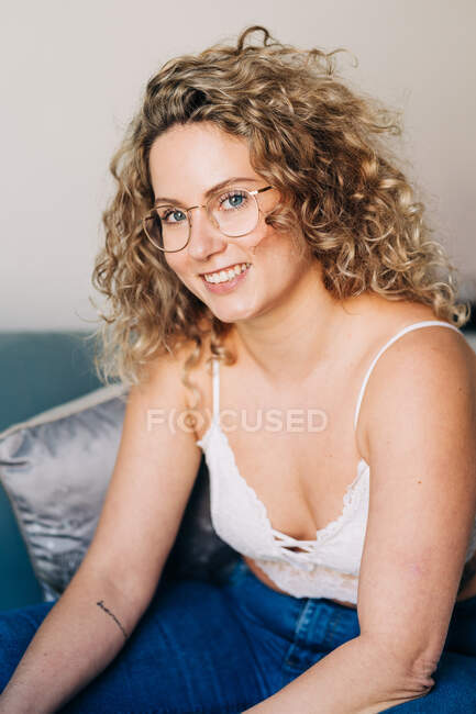 High angle of confident young lady with curly blond hair in stylish outfit and eyeglasses smiling and looking at camera while sitting on sofa at home — Fotografia de Stock
