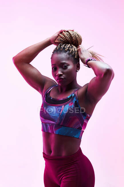 Musculosa atleta afroamericana en ropa deportiva tocando trenzas afro en moño mientras mira la cámara - foto de stock