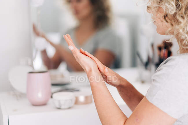 Обрезание женщин в повседневной рубашке, нанося увлажняющий крем для рук, сидя за столом тщеславия в светлой спальне — стоковое фото