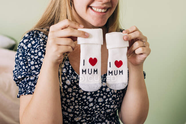 Crop unrecognizable content expectant female in ornamental apparel showing socks with I Mum inscription and heart symbol in house — Stock Photo
