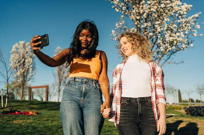 Niedriger Winkel fröhlicher multirassischer Freundinnen, die sich im Frühlingsgarten an den Händen halten und an sonnigen Tagen Selbstaufnahmen mit dem Smartphone machen — Stockfoto