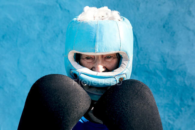 Femme mûre en vêtements de sport et gants de boxe debout avec casque contre le mur bleu et regardant la caméra — Photo de stock