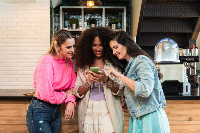 Young stylish female friends browsing mobile phone while standing in the cafeteria — Stock Photo