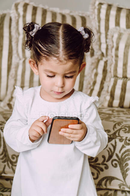 Entzückendes kleines Kind, das in der Nähe der Couch zu Hause steht und interessante Zeichentrickfilme auf dem Handy anschaut — Stockfoto