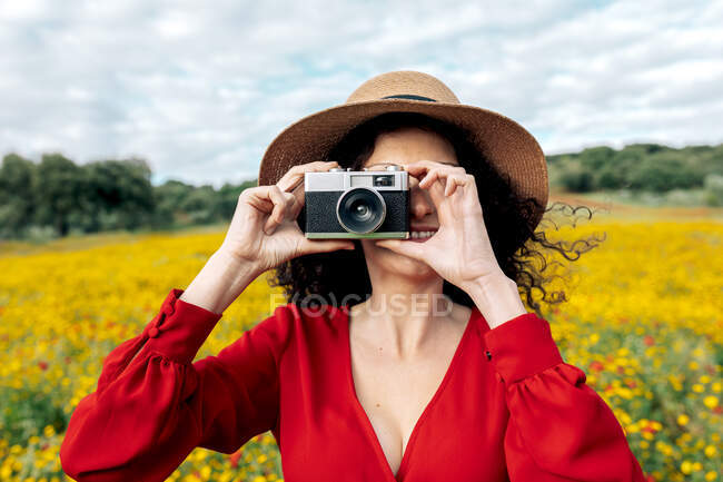 Анонимная улыбающаяся женщина в шляпе фотографирует на винтажную камеру на лугу под облачным небом — стоковое фото