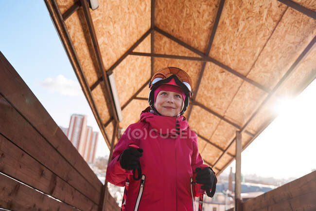 Niña alegre con casco de esquí rosa y ropa deportiva caliente de pie en el club deportivo al aire libre soleado y mirando hacia otro lado con sonrisa - foto de stock