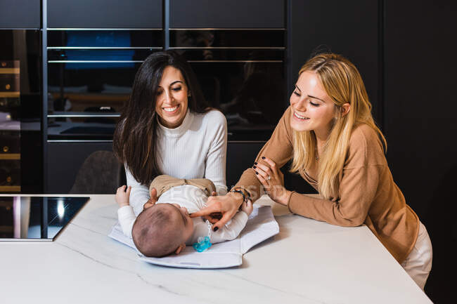 Cheerful couple of lesbian women caressing cute toddler lying on counter in kitchen — Stock Photo