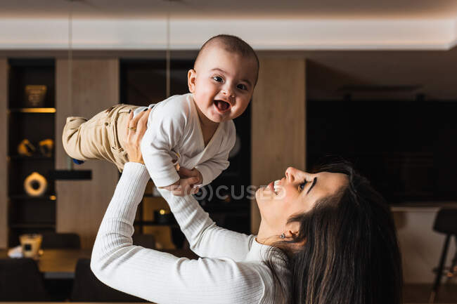 Вид сбоку на восхищенную мать, восхитительно улыбающуюся малышку и веселящуюся вместе дома — стоковое фото