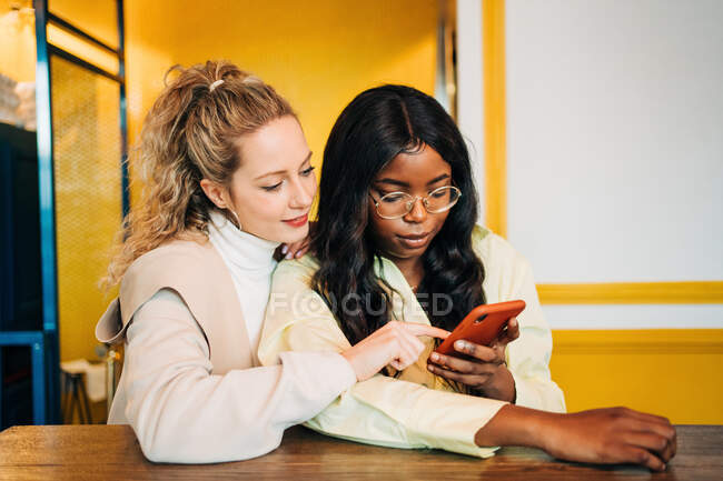 Deleitado multirracial femenina mejores amigos sentados en la cafetería y la navegación en el teléfono inteligente mientras disfrutan de fin de semana juntos - foto de stock