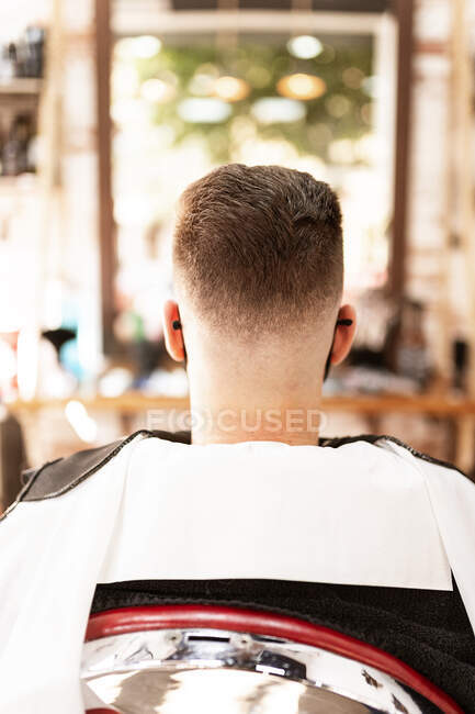 Back view of anonymous male with brown hair in cape sitting in hairdressing salon on blurred background — Fotografia de Stock