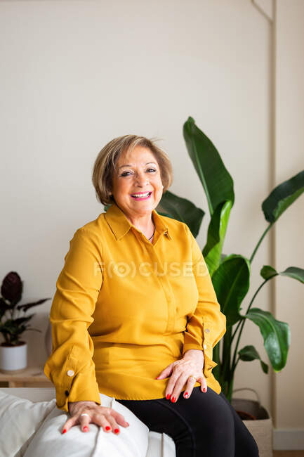 Entzückte reife Frau sitzt auf dem Sofa und blickt in die Kamera im gemütlichen Wohnzimmer mit Zimmerpflanzen — Stockfoto