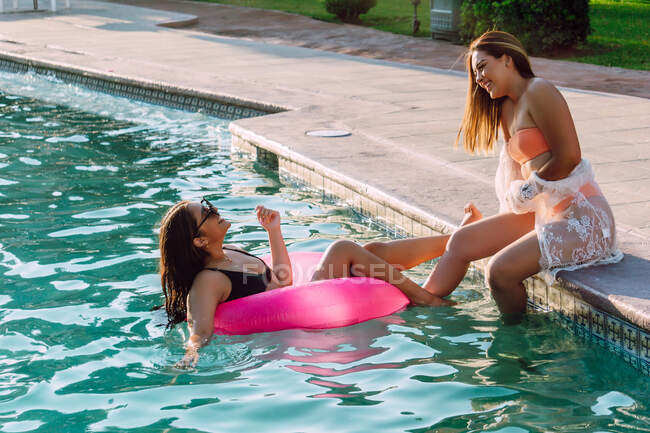 Side view of smiling female traveler in rubber ring holding hand of cheerful friend on poolside during vacation — Stock Photo