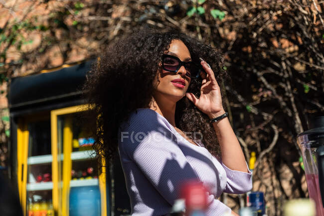 На вид прекрасна молода жінка торкається стилізованих сонцезахисних окулярів і дивиться на камеру, стоячи на сонячному подвір'ї. — стокове фото