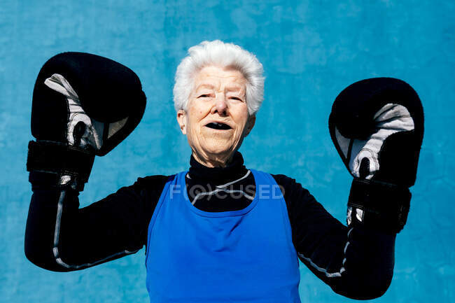 Femme âgée gaie en vêtements de sport levant les mains dans des gants de boxe contre le mur bleu et regardant la caméra — Photo de stock