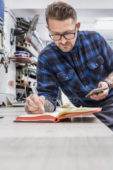 Konzentrierter bärtiger Handwerker steht an der Werkbank und schreibt in Notizblock, während er das Smartphone in der Werkstatt benutzt — Stockfoto
