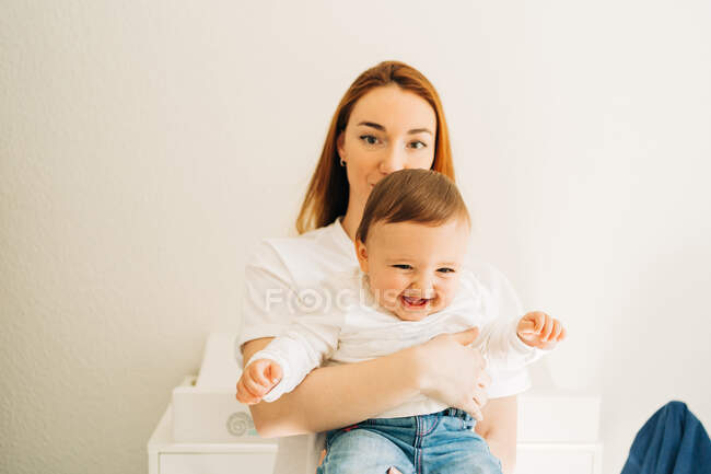 Молода мати в повсякденному одязі дивиться на камеру, тримаючи маленьку дитину в світлій кімнаті — стокове фото