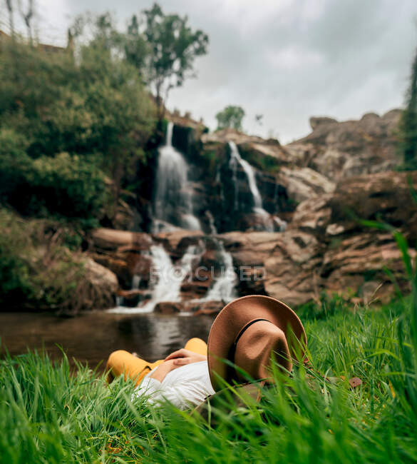 Voyageur masculin anonyme en chapeau relaxant au bord du lac luxuriant et bénéficiant d'une vue rapide en cascade dans la nature estivale — Photo de stock