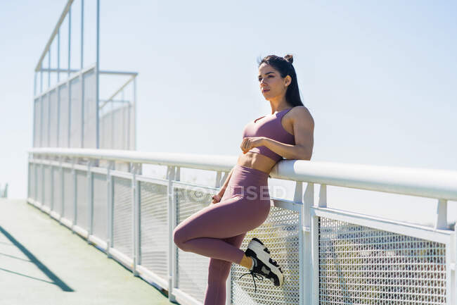 Vista lateral de la joven atleta deportiva con la pierna levantada mirando hacia otro lado en el puente a la luz del sol - foto de stock