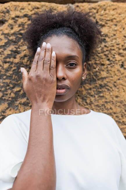 Emotionless joven afroamericana mujer en camisa blanca cubriendo media cara con la mano y mirando a la cámara contra la pared desigual áspera - foto de stock