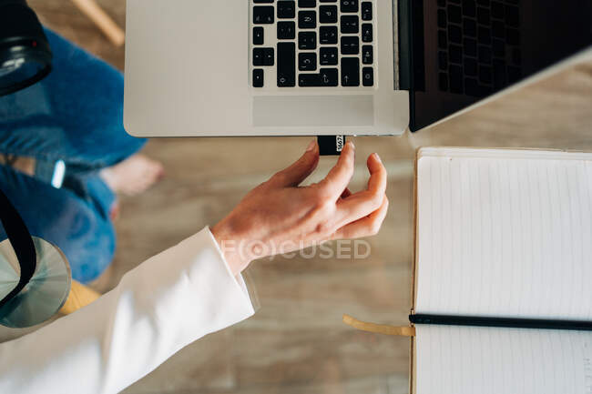 Top view of crop unrecognizable female photographer inserting memory card of photo camera into laptop sitting at glass table — Stock Photo