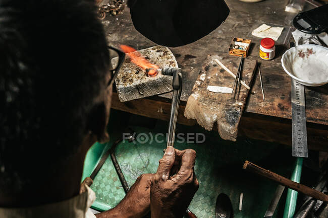 From above cropped unrecognizable of mature ethnic man with burning torch heating piece of metal working at desk in craft workshop — Stock Photo