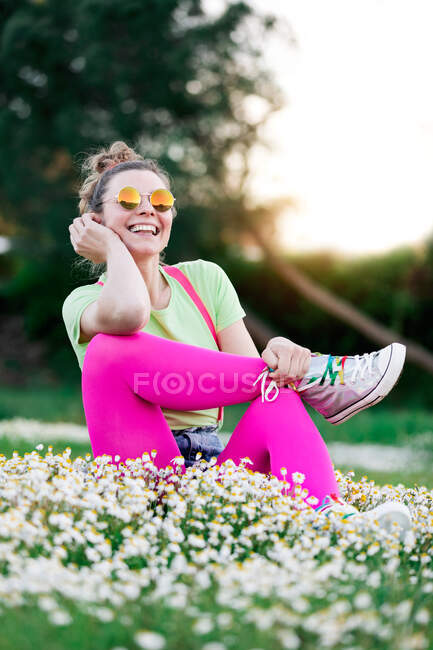 Cheerful young female wearing colorful bright outfit sitting on verdant grassy lawn in summer nature with toothy smile — Stock Photo