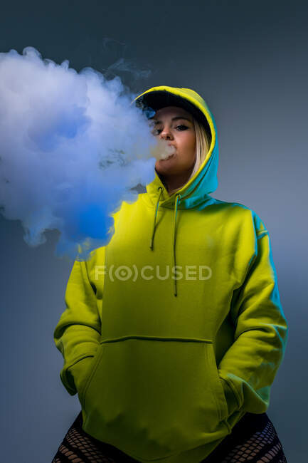 Von unten selbstbewusste Hipsterin im Kapuzenpulli, die im Studio vor grauem Hintergrund E-Zigarette raucht und in die Kamera blickt — Stockfoto