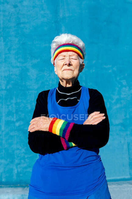 Крутая пожилая женщина в повязке и браслетах, стоящая со скрещенными руками на синем фоне — стоковое фото
