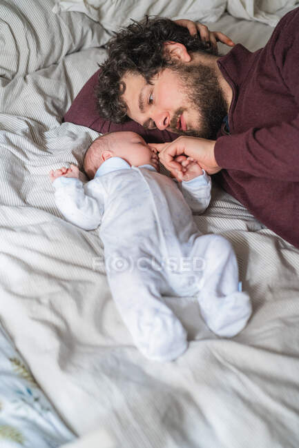 Alto ángulo de papá barbudo abrazando a un niño pequeño y lindo mientras está acostado en la cama arrugada y mirándose el uno al otro - foto de stock