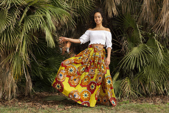 Cheerful young ethnic female in ornamental African skirt looking at camera against palm plants on meadow — Stock Photo