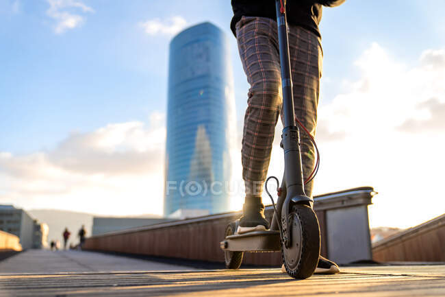 Cultivé entrepreneur ethnique masculin méconnaissable équitation scooter électrique sur passerelle de pont de la ville contre les bâtiments sous ciel bleu nuageux — Photo de stock