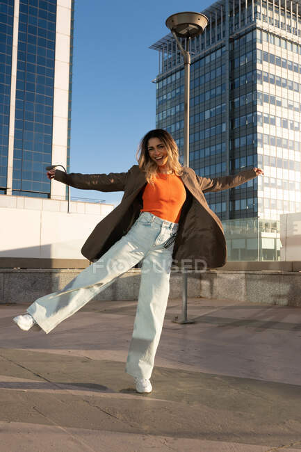 Content female in trendy wear standing with raised leg and outstretched arms while looking at camera in city — Stock Photo
