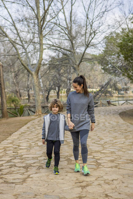 Madre en ropa deportiva sosteniendo al niño a mano mientras pasea por la pasarela y habla contra los árboles - foto de stock