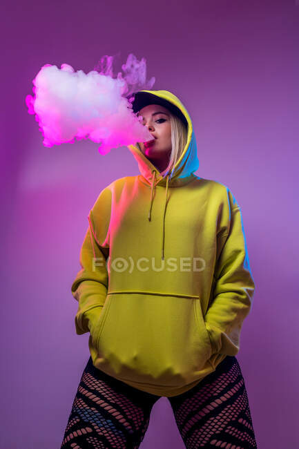 Знизу впевнена жінка-хіпстер у светрі курить електронну сигарету в студії на рожевому фоні, дивлячись на камеру — стокове фото