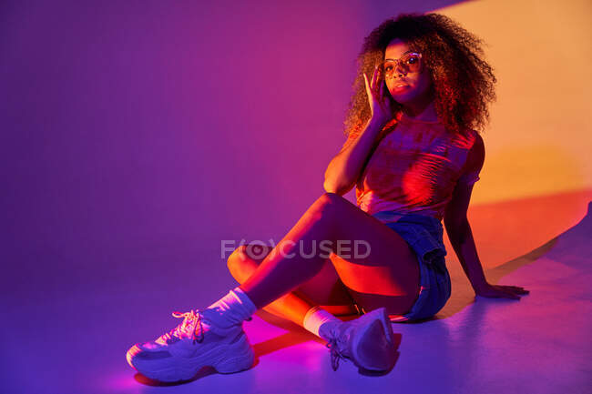 Corpo pieno giovane attraente donna afroamericana in pantaloncini e occhiali da sole alla moda seduto sul pavimento in luci al neon e abbracciare le ginocchia mentre guarda la fotocamera — Foto stock