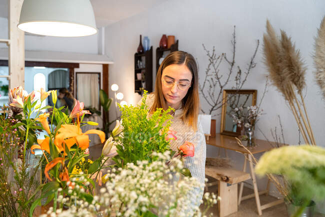 Концентрований молодий жіночий флорист в фартухах і окулярах організовує ароматні жовті квіти у вазі під час роботи в квітковому магазині — стокове фото
