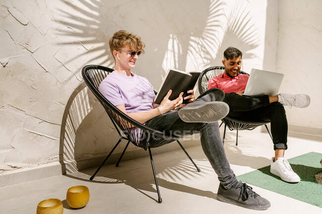 Ganzkörper stilvolle junge multiethnische männliche Freunde mit Laptop und Lesebuch, während sie es sich an sonnigen Tagen auf Stühlen auf der Terrasse gemütlich machen — Stockfoto