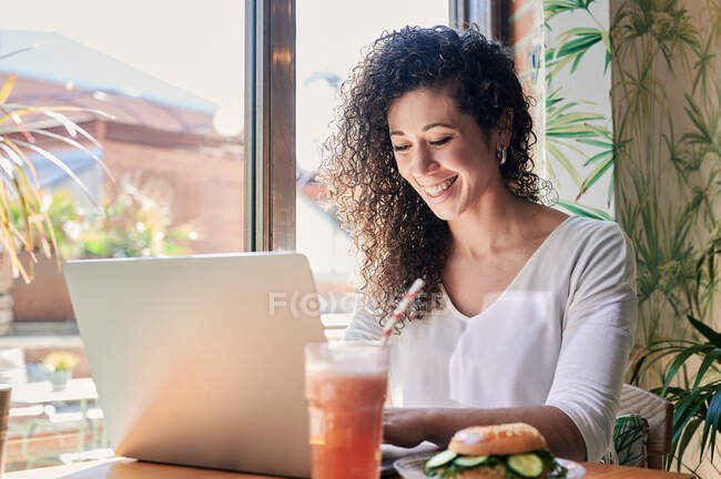 Vista lateral da feliz funcionária étnica trabalhando no netbook à mesa com delicioso sanduíche de bagel vegetariano e bebidas — Fotografia de Stock