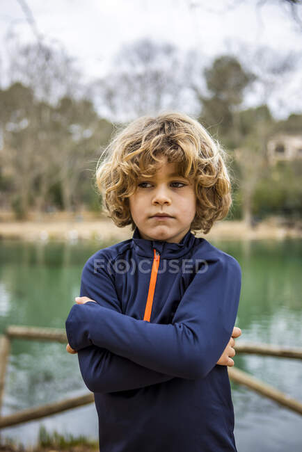 Lonely child in blue clothes with folded arms looking away against water in daylight — Stock Photo