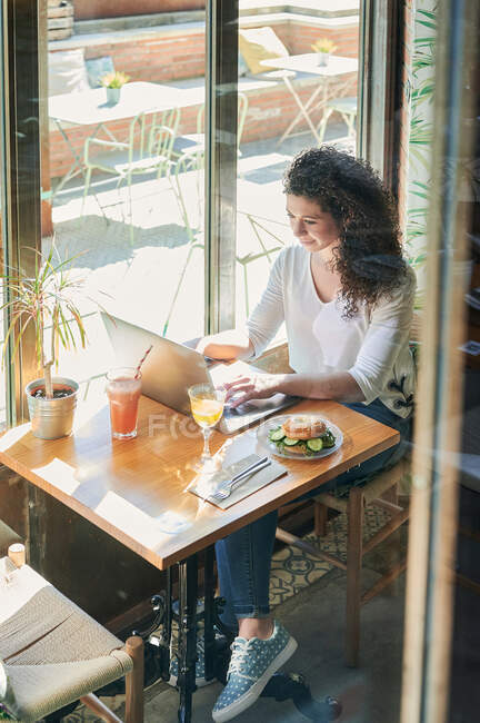 Dall'alto attraverso la vista di vetro di dipendente etnica femminile che lavora su netbook a tavola con delizioso panino vegetariano bagel e bevande — Foto stock