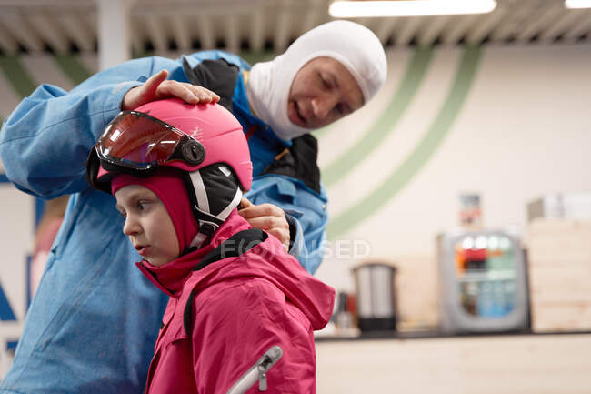 Positive young father in warm activewear and ski balaclava putting protective helmet on cute daughter head in modern ski center — Stock Photo
