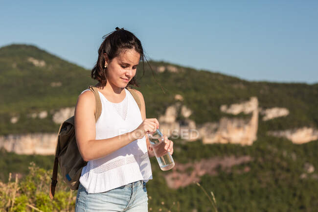 Vista laterale dell'escursionista con zaino aprendo una bottiglia d'acqua rinfrescante durante il trekking in montagna in estate — Foto stock